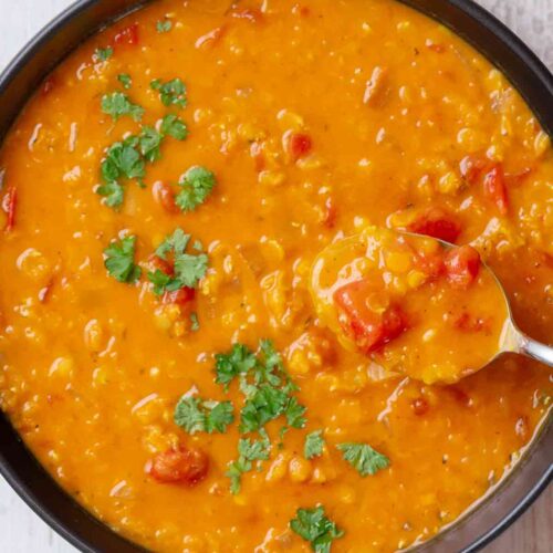 Eine Schüssel mit cremiger roter Linsensuppe mit Kokosmilch, Tomatenstücken und frischer Petersilie. Ein Löffel mit Suppe wird aus der Schüssel gehoben.