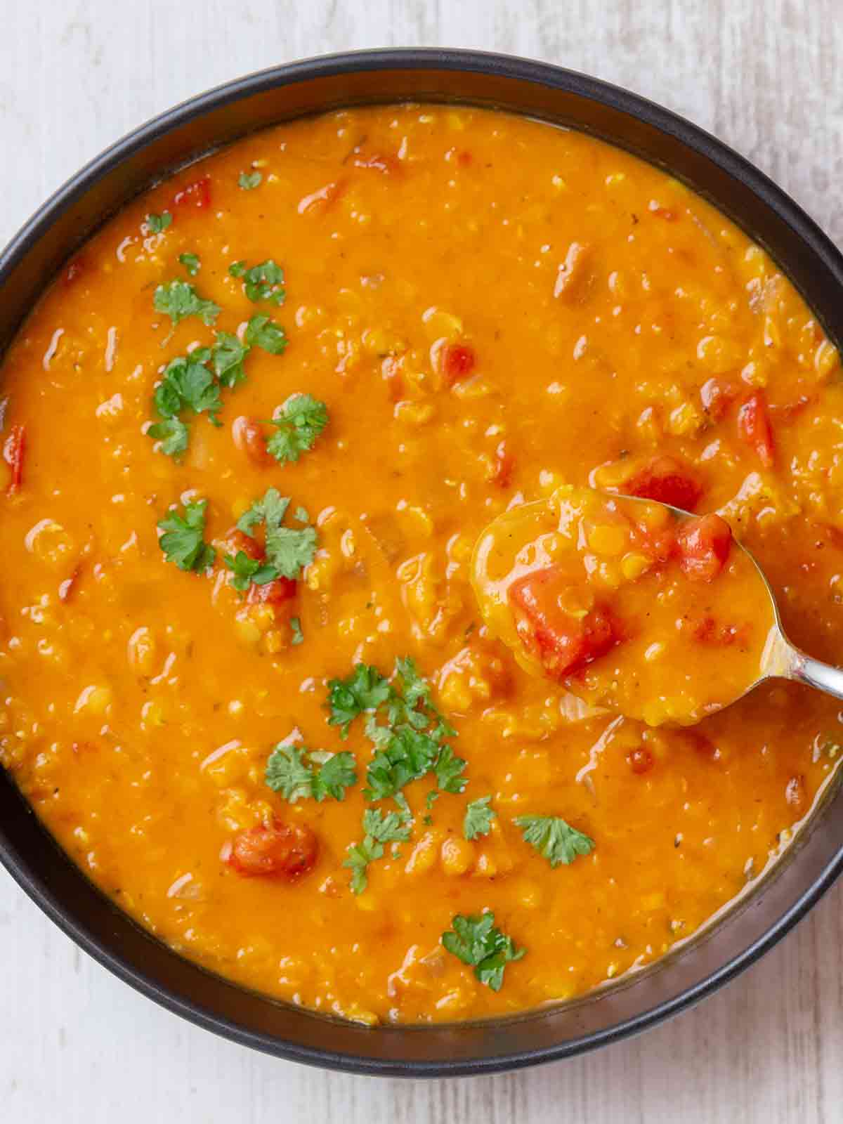 Eine Schüssel mit cremiger roter Linsensuppe mit Kokosmilch, Tomatenstücken und frischer Petersilie. Ein Löffel mit Suppe wird aus der Schüssel gehoben.