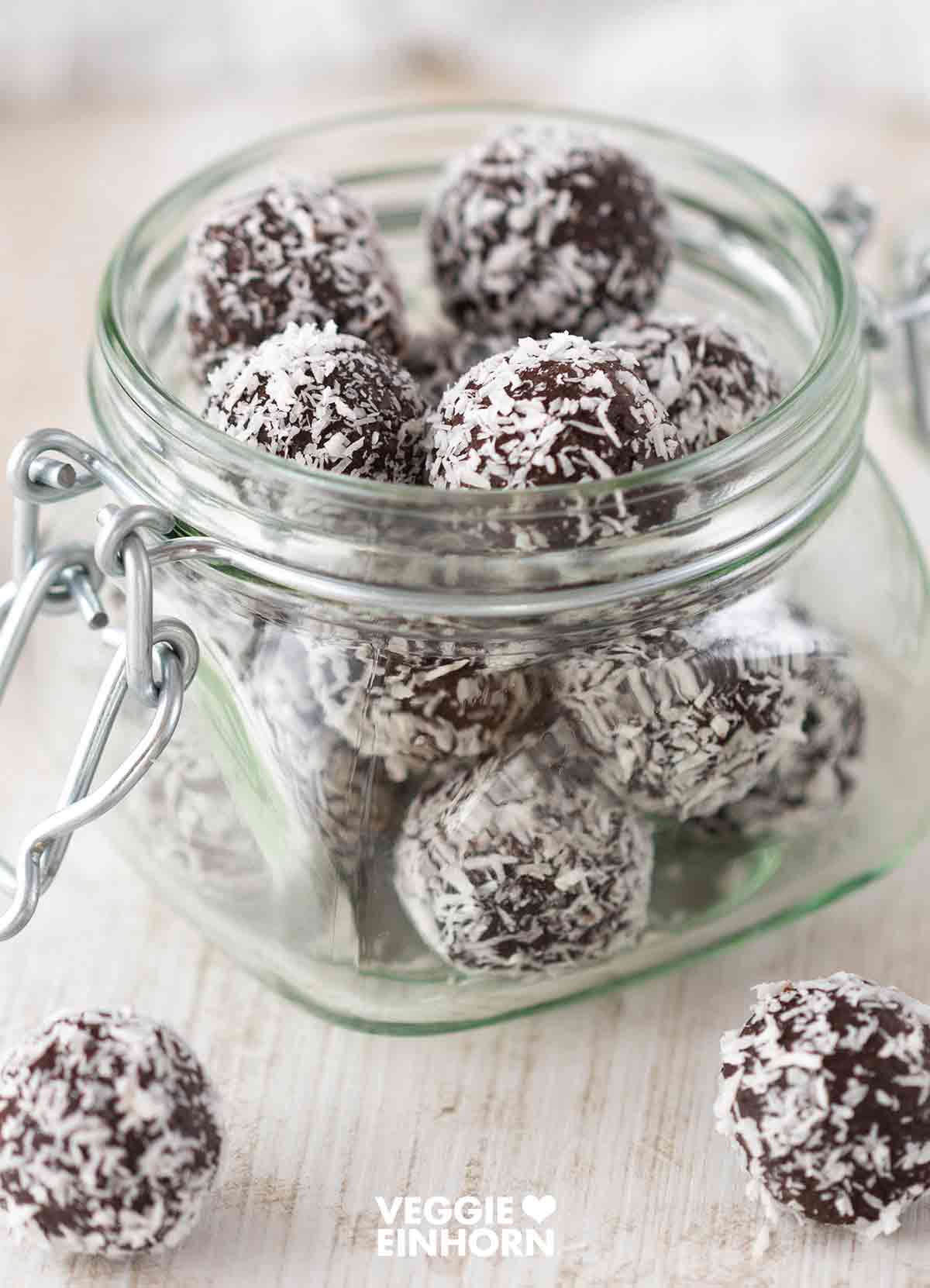 Ein geöffnetes Glas mit dunklen veganen Dattel-Energiebällchen, die mit Kokosraspeln bestreut sind. Einige der Kugeln liegen auf einem hellen Holztisch verstreut.