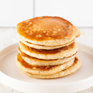 Ein Stapel goldbrauner, fluffiger veganer Pancakes auf einem weißen Teller, vor einem hellen Hintergrund.