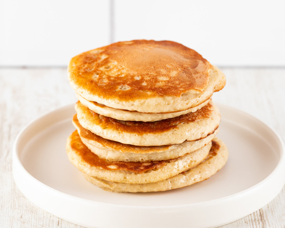Ein Stapel goldbrauner, fluffiger veganer Pancakes auf einem weißen Teller, vor einem hellen Hintergrund.