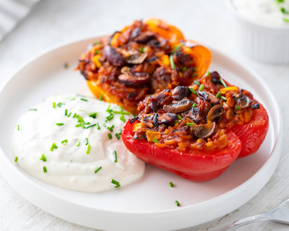 Vegane gefüllte Paprika mit einer herzhaften Mischung aus Reis, Kidneybohnen, Mais und Champignons, serviert auf einem weißen Teller mit veganem Kräuterquark.