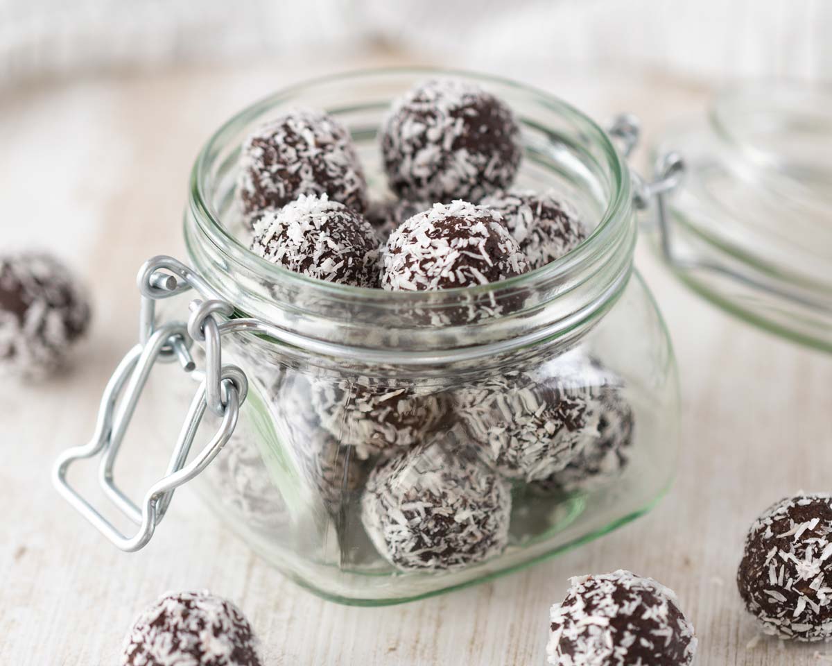 Ein geöffnetes Glas mit veganen Energiekugeln, die mit Kokosraspeln bestreut sind. Einige der Kugeln liegen auf einem hellen Holztisch um das Glas herum.