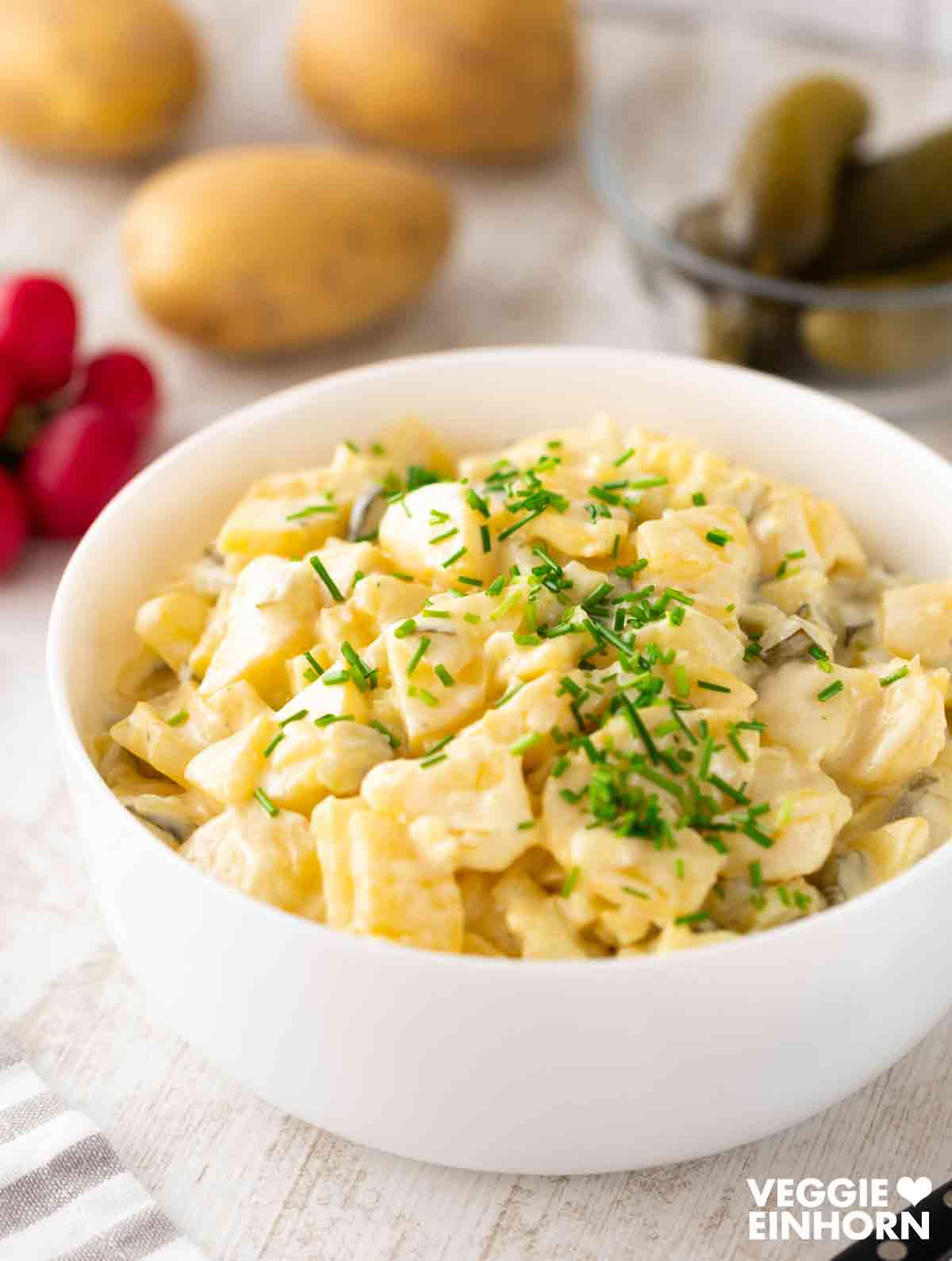 Vegane Kartoffelsalat mit Mayonnaise in einer weißen Schüssel, garniert mit frischem Schnittlauch; im Hintergrund liegen ganze Kartoffeln, Radieschen und Gewürzgurken.