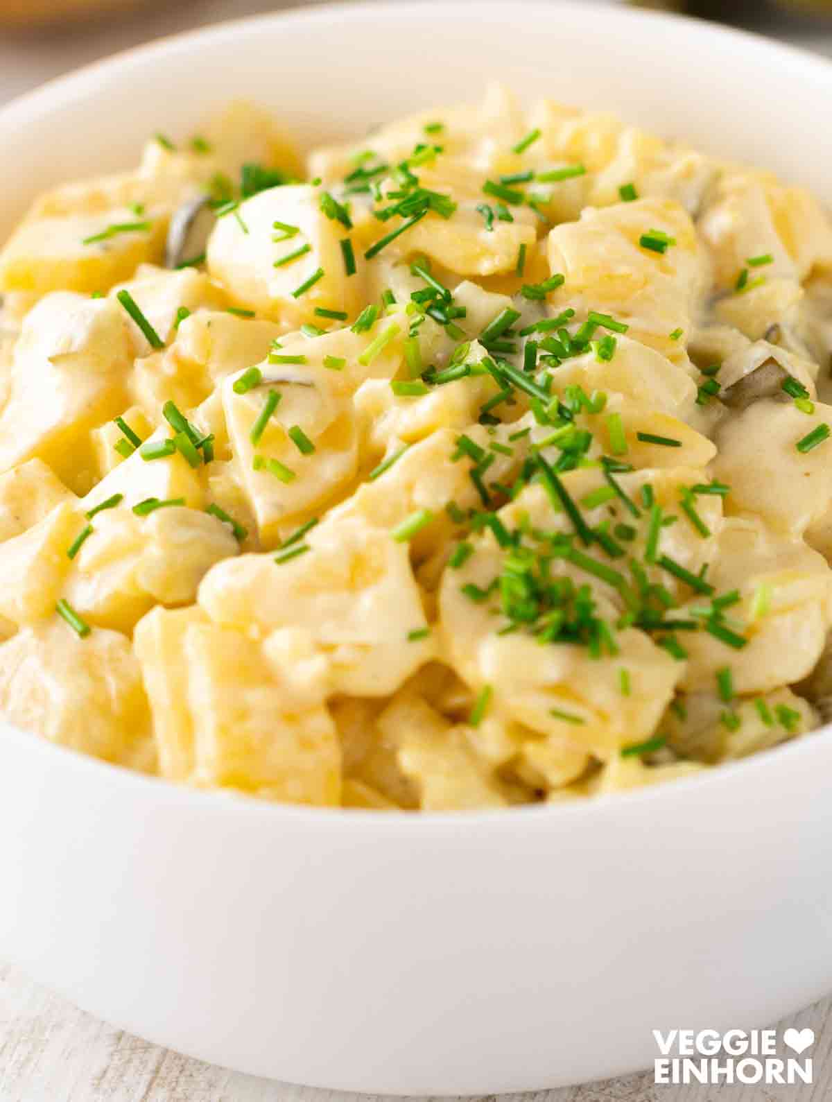 Cremiger veganer Kartoffelsalat mit veganer Mayonnaise und Schnittlauchringen, serviert in einer weißen Schale.