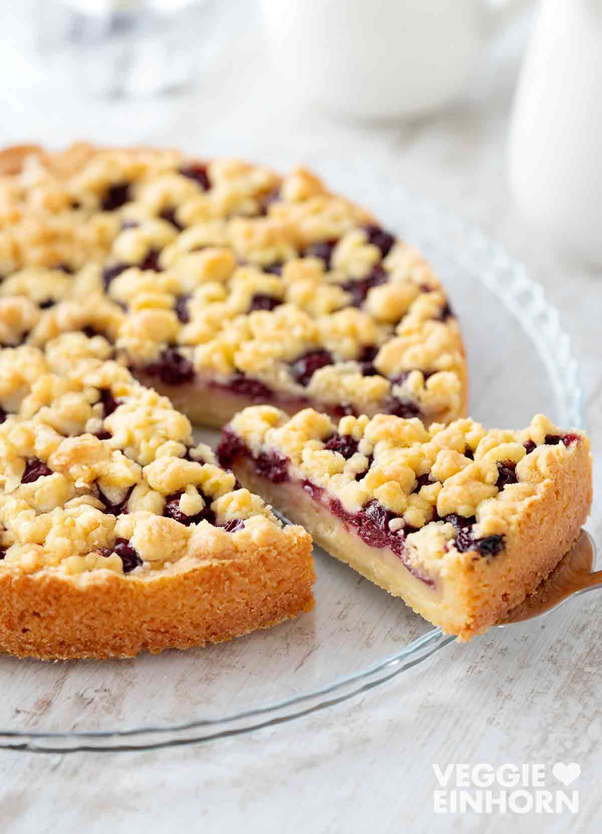 Ein veganer Kirschkuchen mit Pudding und Streuseln auf einem gläsernen Kuchenteller. Ein Stück wurde herausgehoben, sodass die vegane Puddingfüllung, die saftigen Kirschen und die knusprigen Streusel gut sichtbar sind. Im Hintergrund stehen eine weiße Milchkanne und ein Glas Wasser. 