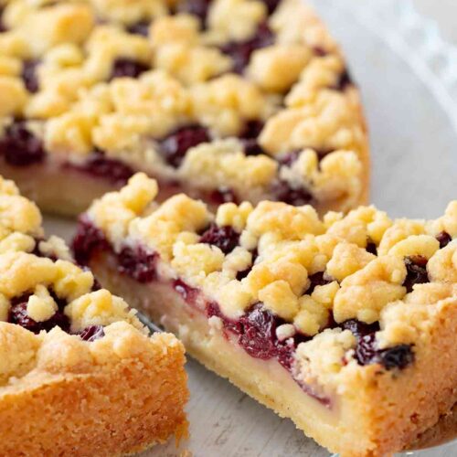 Nahaufnahme eines veganen Kirsch-Streuselkuchens mit einer Schicht Pudding auf einem gläsernen Kuchenteller. Ein Stück wurde herausgeschnitten.