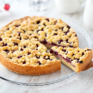 Ein veganer Streuselkuchen mit Kirschen und Puddingfüllung auf einem gläsernen Kuchenteller. Ein Stück wurde herausgeschnitten und liegt auf einem Kuchenheber.