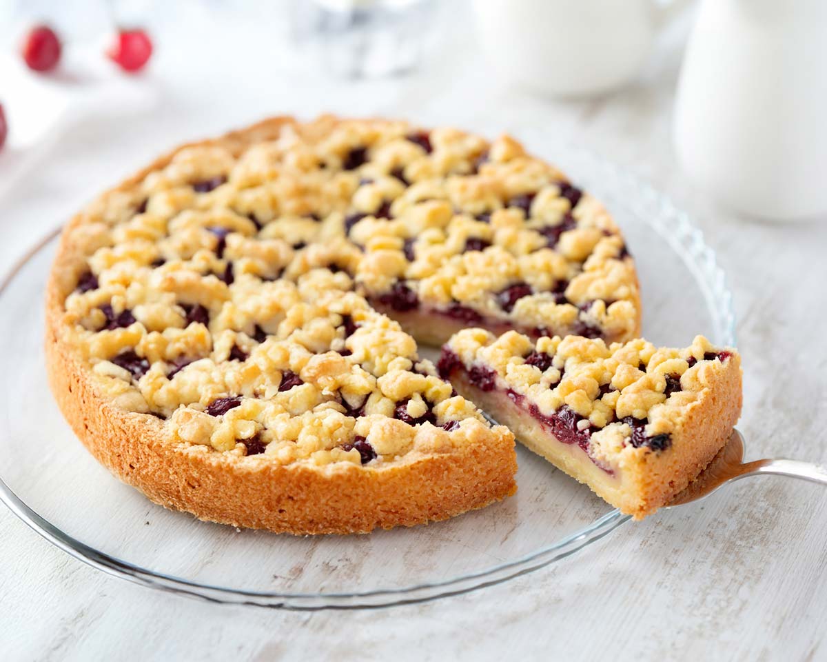 Ein veganer Streuselkuchen mit Kirschen und Puddingfüllung auf einem gläsernen Kuchenteller. Ein Stück wurde herausgeschnitten und liegt auf einem Kuchenheber.