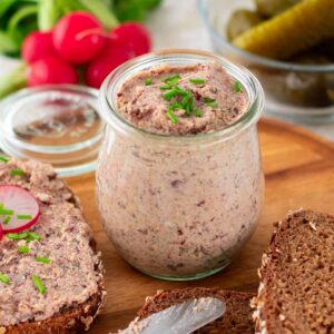 Ein Glas mit veganer Leberwurst, garniert mit frischem Schnittlauch, steht auf einem runden Holzbrett. Daneben eine Scheibe Vollkornbrot mit dem Aufstrich, dekoriert mit Radieschenscheiben und Schnittlauch. Im Hintergrund eine Schale mit Gewürzgurken und ein Bund frische Radieschen.