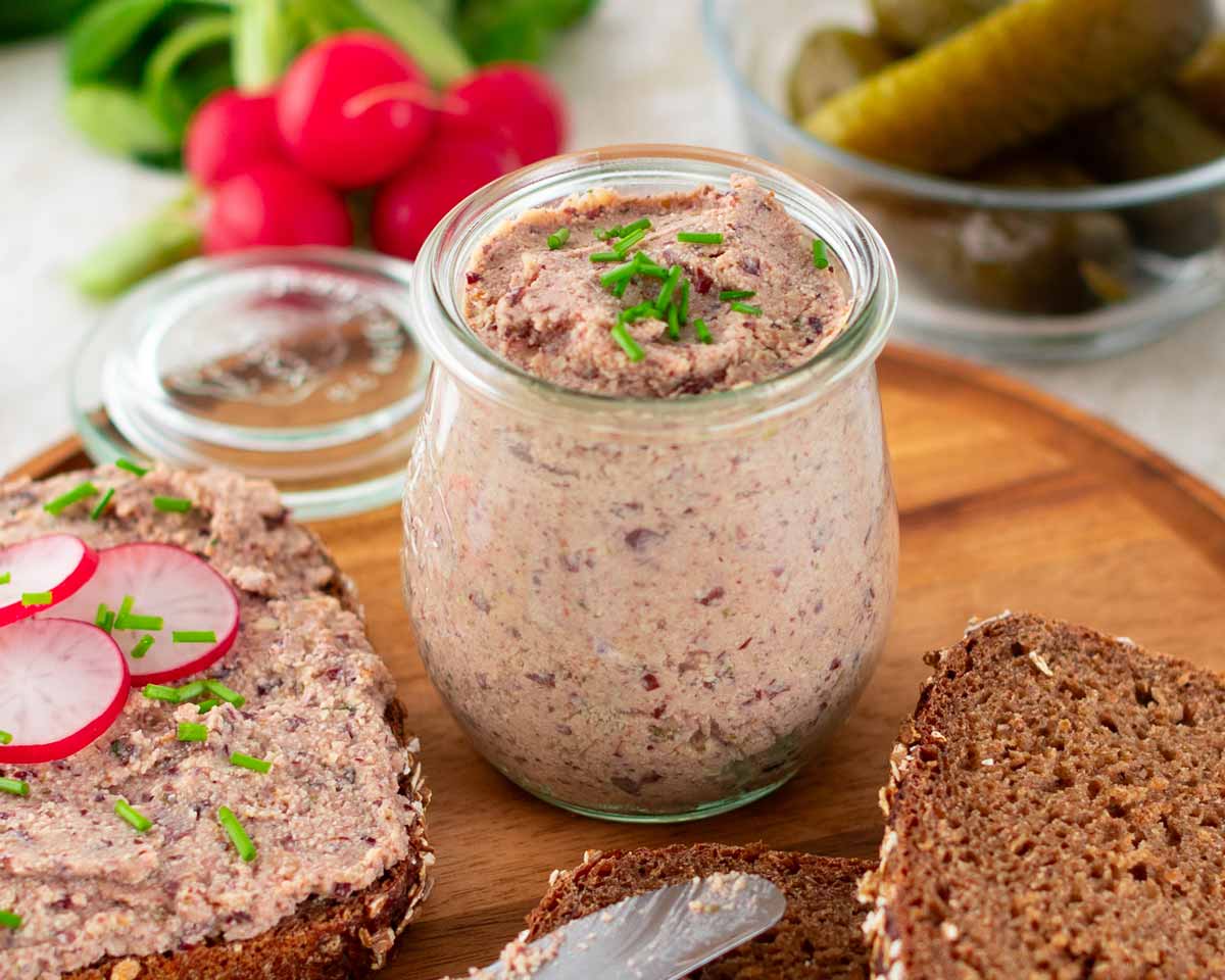 Ein Glas mit veganer Leberwurst, garniert mit frischem Schnittlauch, steht auf einem runden Holzbrett. Daneben eine Scheibe Vollkornbrot mit dem Aufstrich, dekoriert mit Radieschenscheiben und Schnittlauch. Im Hintergrund eine Schale mit Gewürzgurken und ein Bund frische Radieschen.