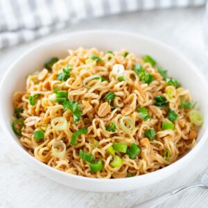 Eine weiße Schüssel mit asiatischen Nudeln in Erdnusssoße, garniert mit Frühlingszwiebeln, gehackten Erdnüssen, Sesam und frischer Petersilie. Im Hintergrund ein Glas Wasser und ein gestreiftes Küchentuch, im Vordergrund eine Gabel auf einem weißen Holztisch.