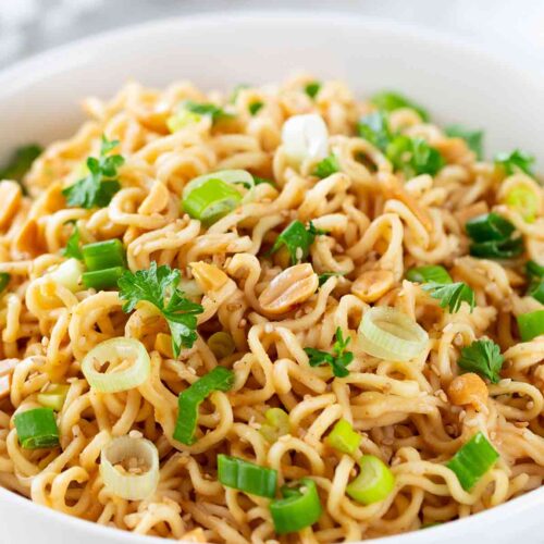 Eine weiße Schüssel mit asiatischen Nudeln in Erdnusssoße, bestreut mit Frühlingszwiebeln, gehackten Erdnüssen, Sesam und frischer Petersilie.