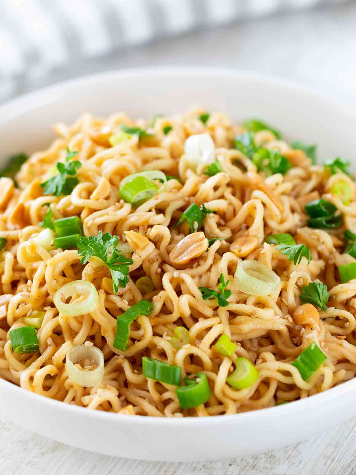 Eine weiße Schüssel mit asiatischen Nudeln in Erdnusssoße, bestreut mit Frühlingszwiebeln, gehackten Erdnüssen, Sesam und frischer Petersilie.