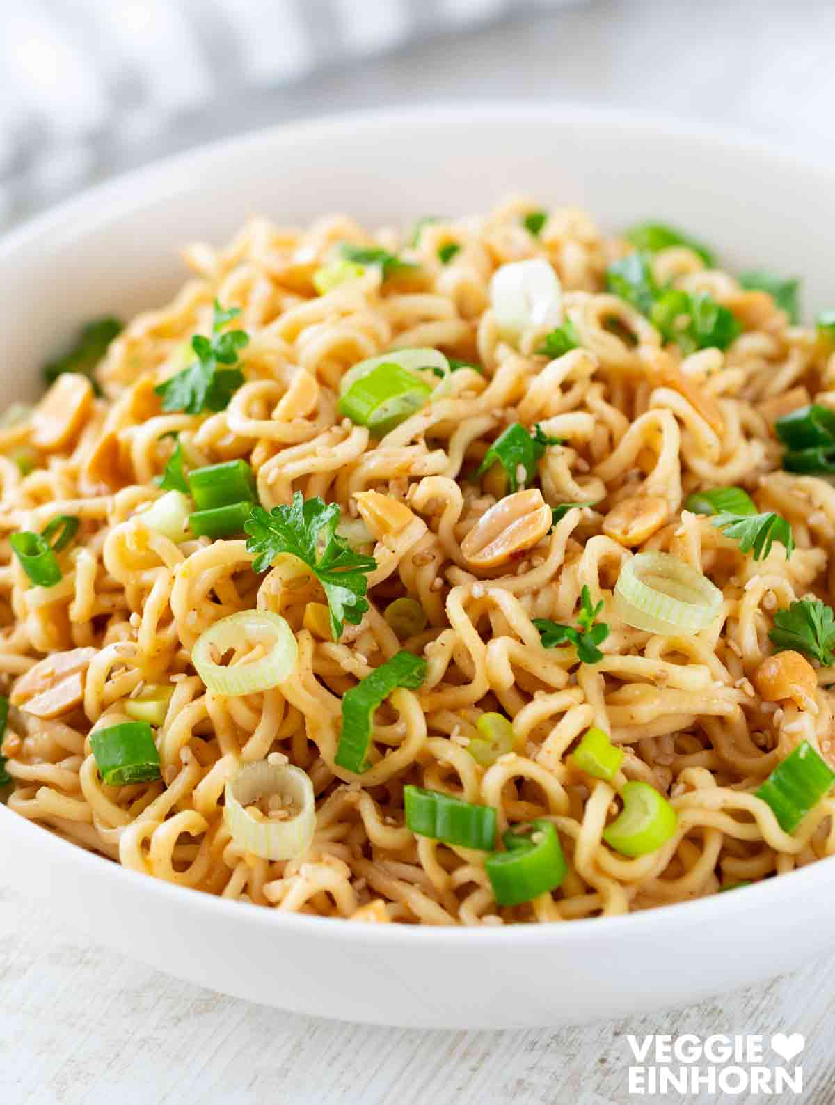 Eine weiße Schüssel mit Mie Nudeln in Erdnusssoße, garniert mit Frühlingszwiebeln, gehackten Erdnüssen, Sesam und frischer Petersilie.