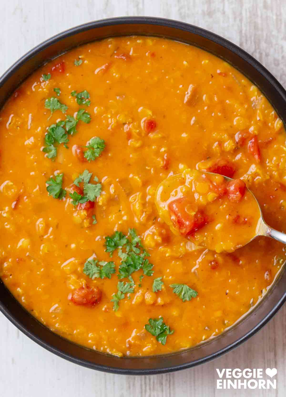 Vegane rote Linsensuppe Eine schwarze Schüssel mit cremiger roter Linsensuppe mit Kokosmilch, garniert mit Petersilie. Ein Löffel mit Suppe wird aus der Schüssel gehoben.