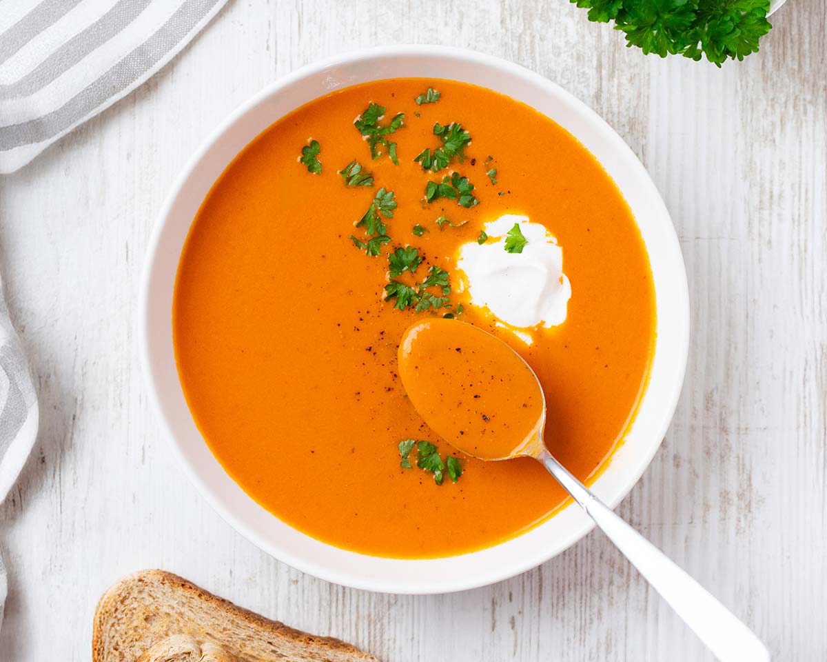 Tomatensuppe mit Kokosmilch auf einem Teller, garniert mit einem Klecks Kokosmilch und frischer Petersilie. Ein Löffel mit Suppe liegt im Teller.