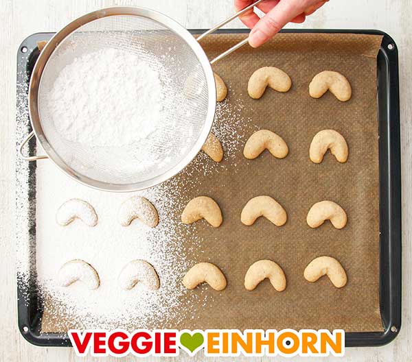 Kipferl mit Puderzucker bestäuben Heiße Vanillekipferl auf dem Backblech werden mit Puderzucker aus einem Sieb bestreut