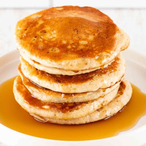 Ein Stapel goldbrauner, fluffiger veganer Pancakes, mit Ahornsirup beträufelt, auf einem weißen Teller vor einem hellen Hintergrund.