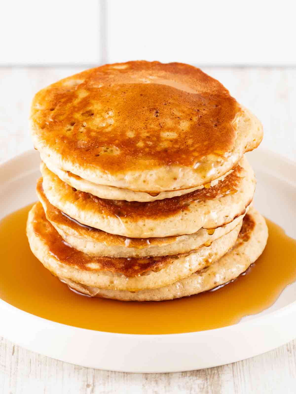 Ein Stapel goldbrauner, fluffiger veganer Pancakes, mit Ahornsirup beträufelt, auf einem weißen Teller vor einem hellen Hintergrund.
