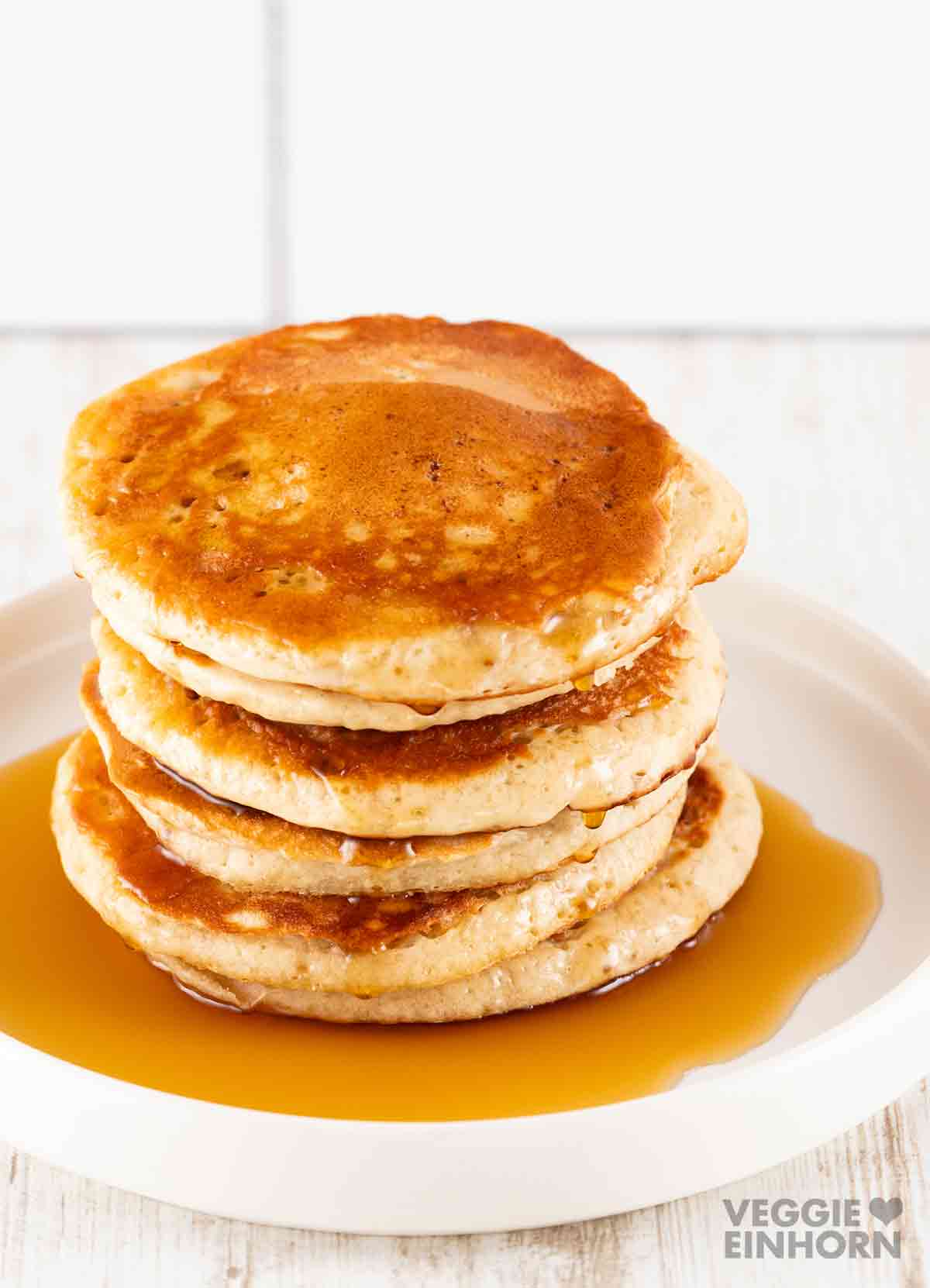 Ein Stapel goldbrauner, fluffiger veganer Pancakes mit Ahornsirup auf einem weißen Teller.