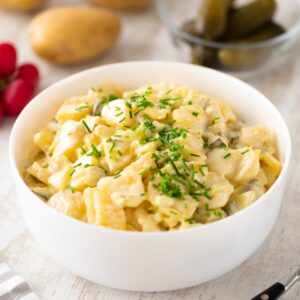Veganer Kartoffelsalat mit veganer Mayonnaise und Schnittlauch in einer weißen Schüssel, im Hintergrund liegen Radieschen, Kartoffeln und eine kleine Glasschale mit Gewürzgurken.