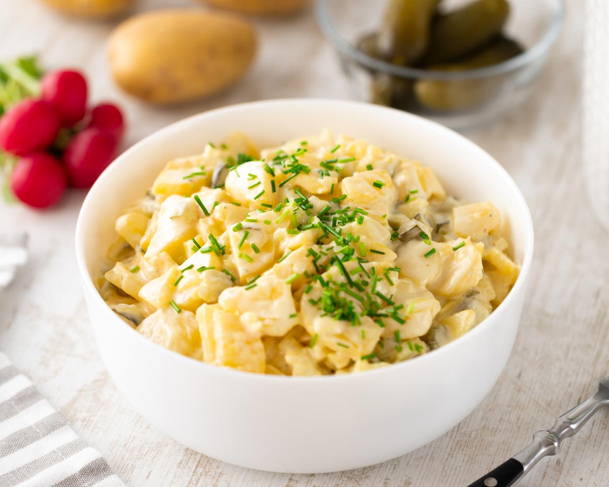 Veganer Kartoffelsalat mit veganer Mayonnaise und Schnittlauch in einer weißen Schüssel, im Hintergrund liegen Radieschen, Kartoffeln und eine kleine Glasschale mit Gewürzgurken.