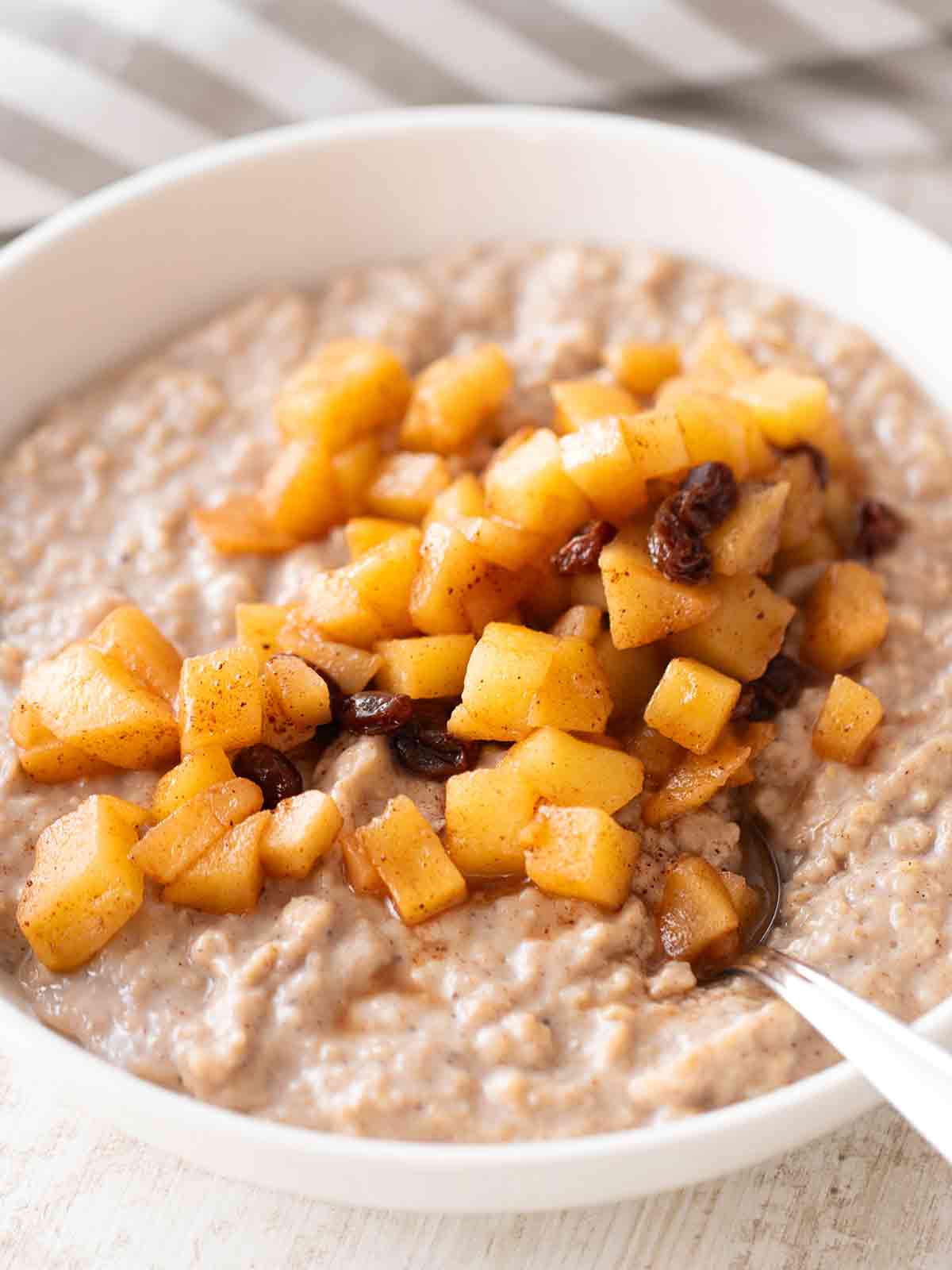 Nahaufnahme von veganem Apfel-Zimt-Porridge mit Apfelstücken und Rosinen in einer weißen Schüssel.