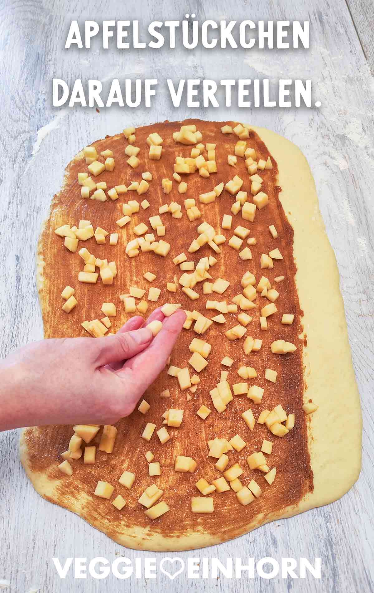 Eine Hand verteilt kleine Apfelstücke auf einem mit Zimt bestrichenen Hefeteig.
