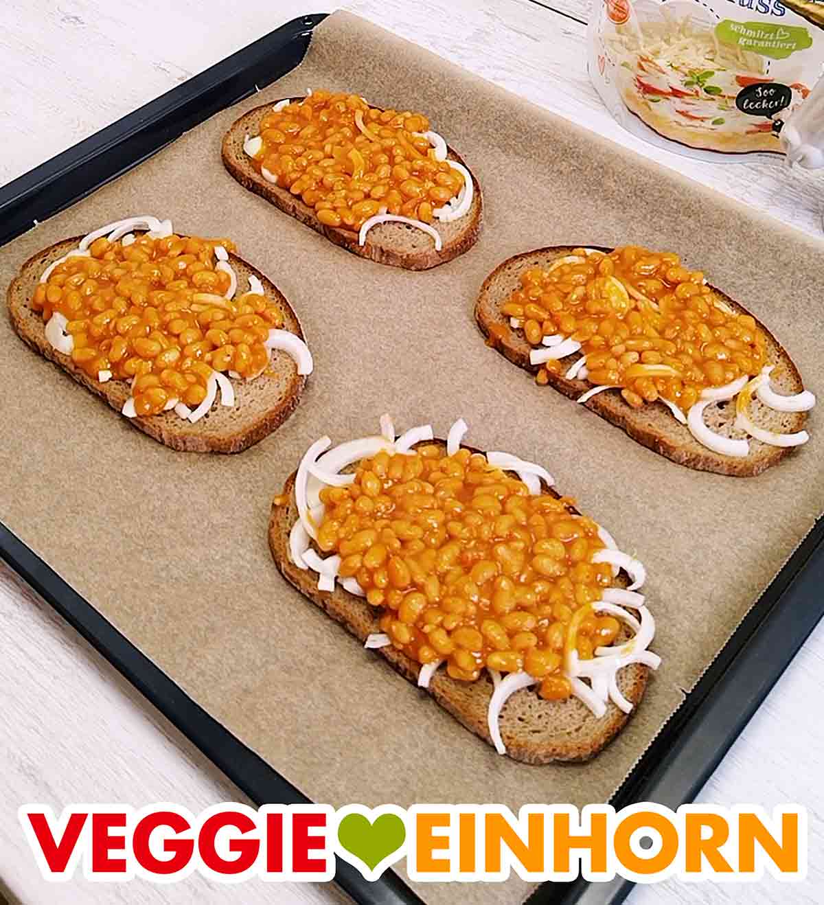 Auf dem mit Backpapier ausgelegten Backblech liegen vier Brotscheiben mit halben Zwiebelringen und Baked Beans. Backblech mit vier Scheiben Brot mit Zwiebeln und Baked Beans
