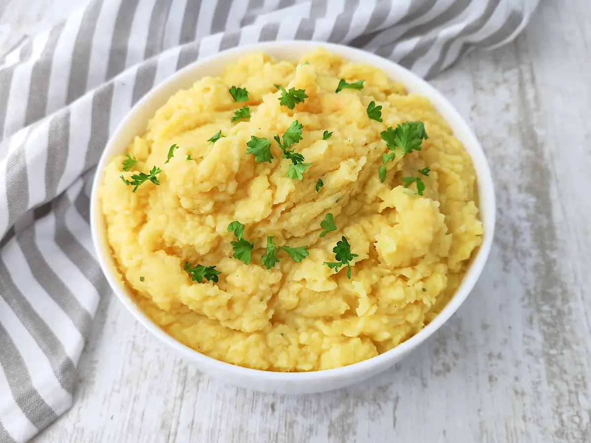 Veganes Kartoffelpüree in einer weißen Schüssel auf einem hellen Holztisch, dekoriert mit frischer Petersilie und einem grau gestreiften Küchentuch im Hintergrund.