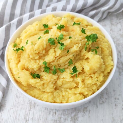 Weiße Schüssel mit cremigem veganem Kartoffelpüree auf hellem Untergrund, garniert mit Petersilie und neben einem grau gestreiften Küchentuch.