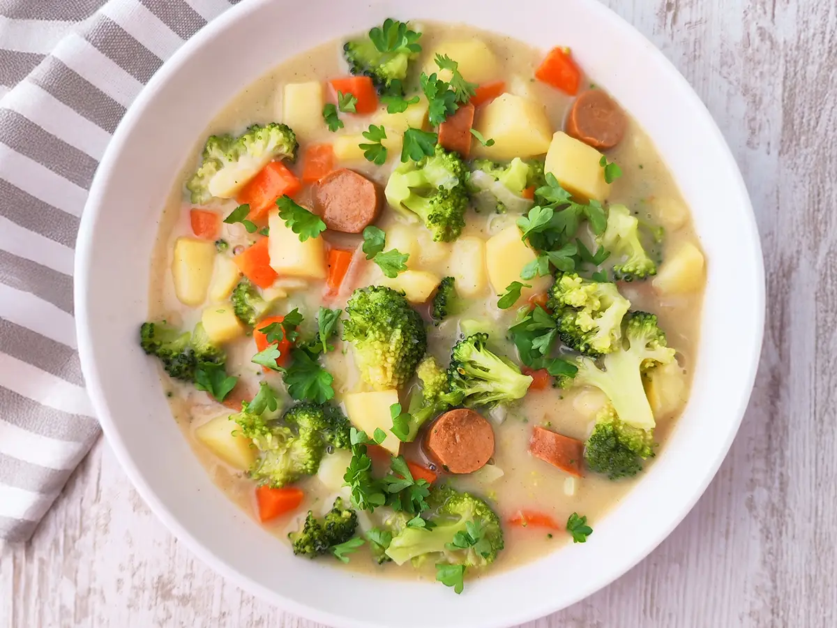 Ein Teller cremiger veganer Gemüseeintopf mit Kartoffeln, Brokkoli, Möhren und veganen Wiener Würstchen, bestreut mit frischer Petersilie.