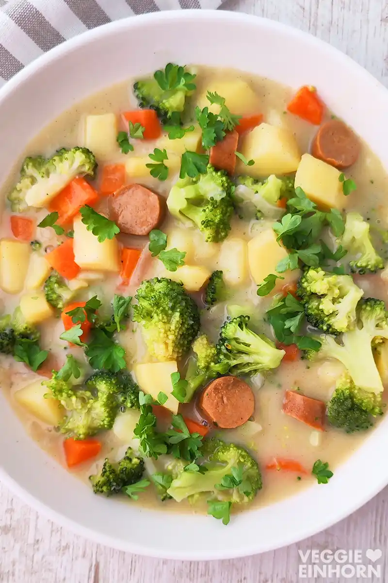 Nahaufnahme von veganem Gemüseeintopf mit Brokkoli, Möhren, Kartoffeln und veganen Wiener Würstchen, bestreut mit frischer Petersilie.