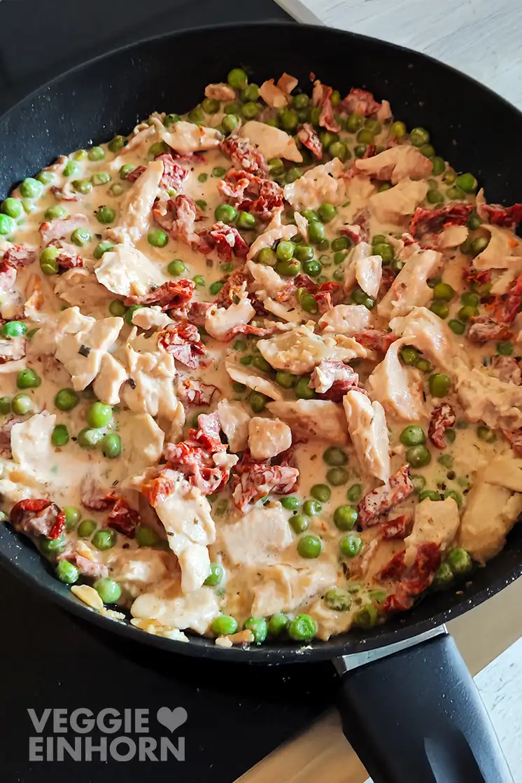 Auf dem Herd stehende schwarze Pfanne mit veganem Hähnchen, getrockneten Tomaten und Erbsen in einer cremigen Soße.