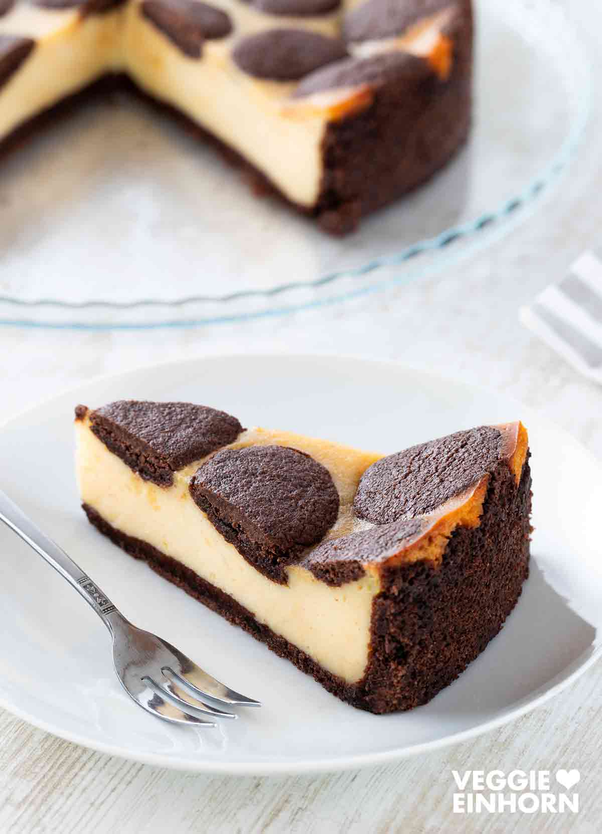 Ein Stück veganer russischer Zupfkuchen mit cremiger Füllung und schokoladigem Mürbeteig auf einem weißen Teller mit einer Kuchengabel, im Hintergrund der angeschnittene Kuchen auf einer Glasplatte.