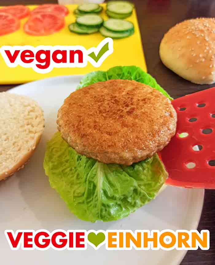 Der vegane Chicken Burger wird zubereitet mit selbst gebackenen Burgerbrötchen, Salat, Tomaten und Gurken. Ein veganer Sieht-Chick-Aus Burger Patty wird auf ein Burgerbrötchen mit einem Salatblatt gelegt.