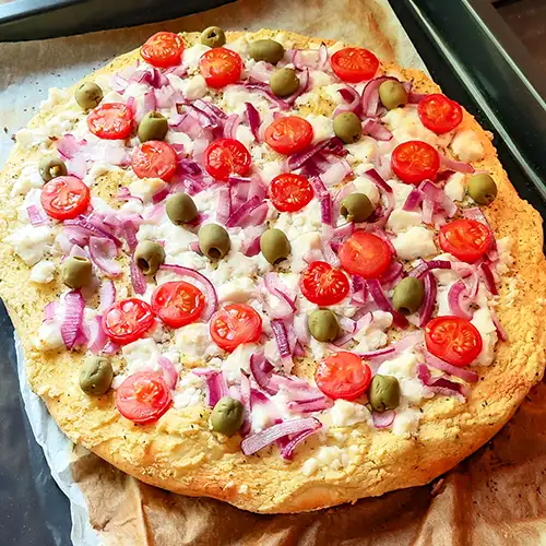 Hummus-Pizza mit veganem Feta, Tomaten, roten Zwiebeln und Oliven auf dem Backblech mit Backpapier.