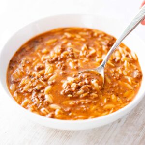 Eine weiße Schüssel mit rötlich-brauner Kritharaki-Suppe, in der kleine Orzo-Nudeln und veganes Hack zu sehen sind. Eine Hand hält einen Löffel, der in die Suppe eingetaucht ist.
