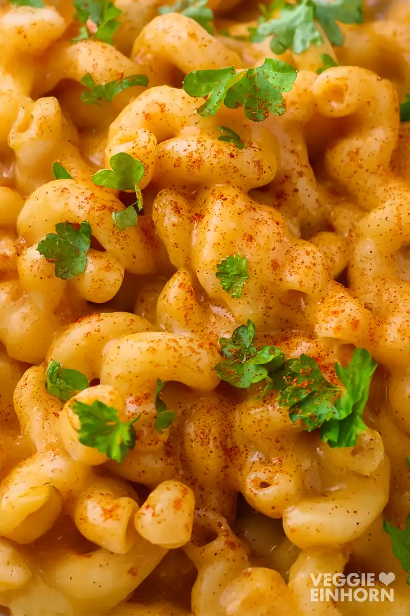 Eine Nahaufnahme von veganen Mac and Cheese, die gleichmäßig mit orangener veganer Käsesoße überzogen und mit Petersilie und Paprikapulver garniert sind.