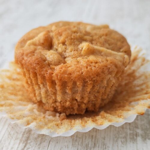 Ein frisch gebackener veganer Apfelmuffin im geöffneten Papierförmchen auf hellem Untergrund.