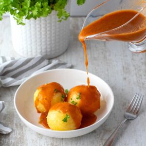 Vegane Bratensoße wird aus einer Sauciere über drei Kartoffelklöße in einem weißen Teller gegossen, garniert mit frischer Petersilie. Im Hintergrund ein Topf mit Petersilie und ein gestreiftes Küchentuch.