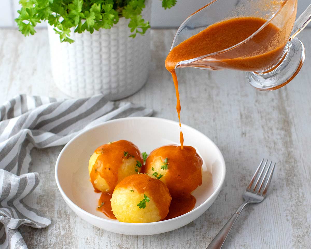 Vegane Bratensoße wird aus einer Sauciere über drei Kartoffelklöße in einem weißen Teller gegossen, garniert mit frischer Petersilie. Im Hintergrund ein Topf mit Petersilie und ein gestreiftes Küchentuch.