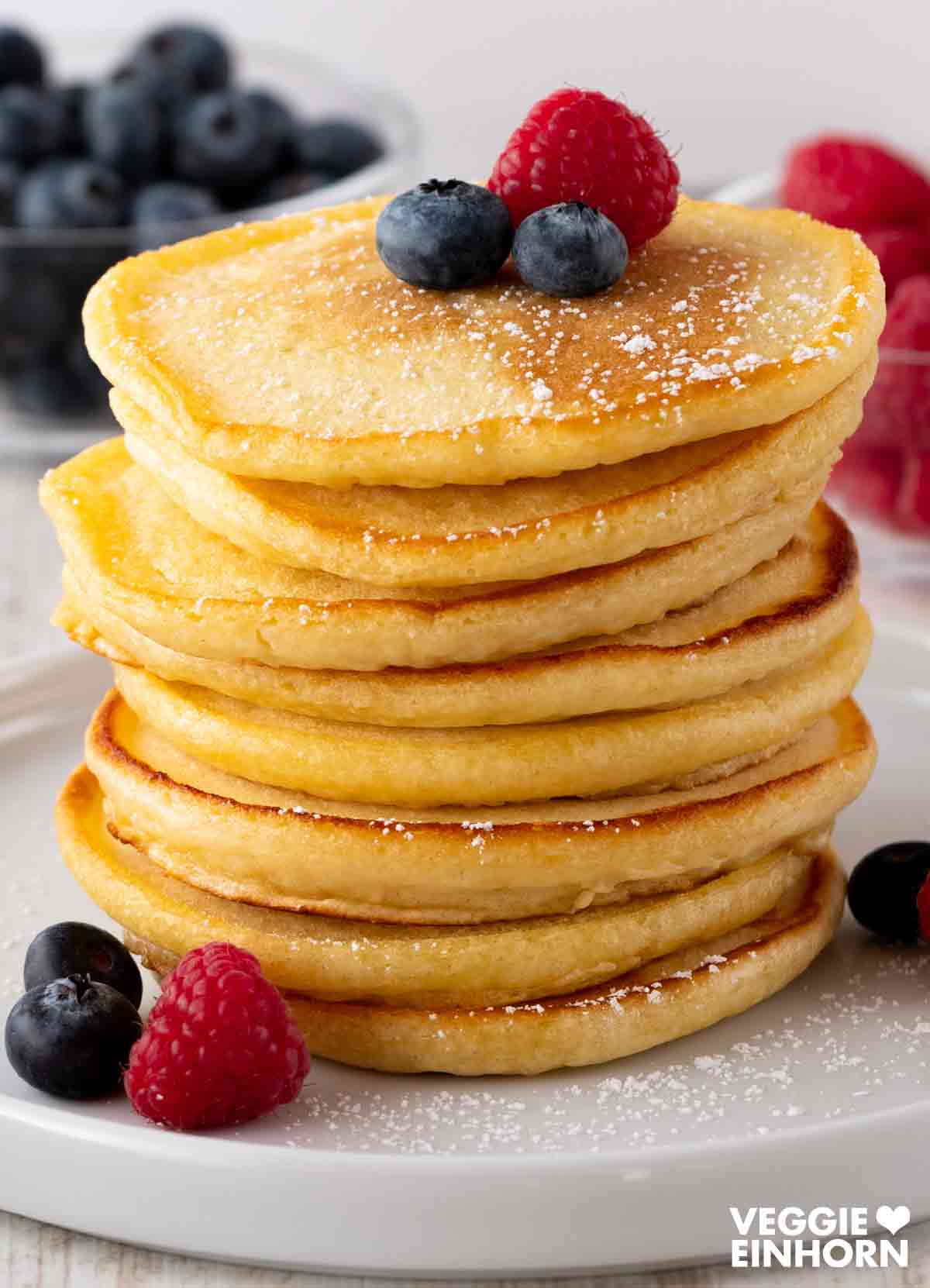 Ein Stapel fluffiger veganer Pancakes, garniert mit Puderzucker und Beeren, auf einem weißen Teller.