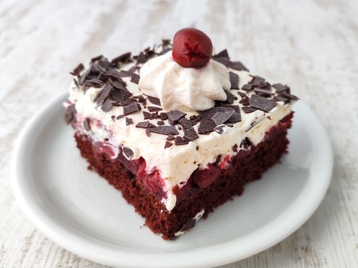 Ein einzelnes Stück vegane Schwarzwälder Kirschtorte vom Blech mit lockerer Schokoschicht, Kirschfüllung, Sahnehaube und Schokoraspeln.