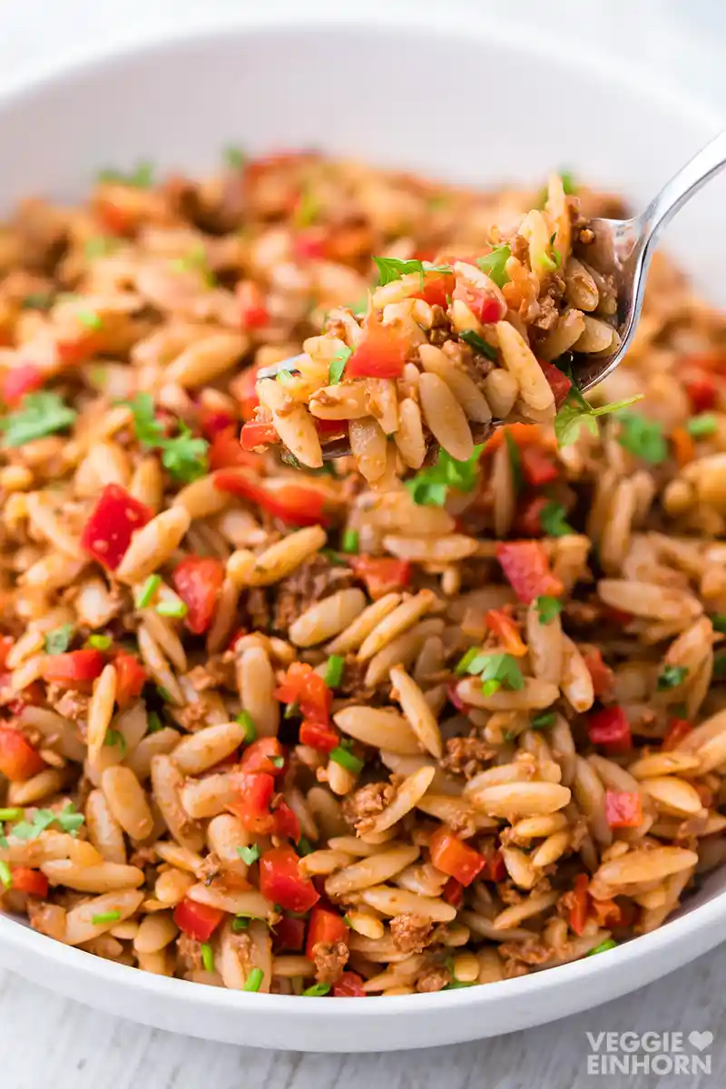 Nahaufnahme eines Kritharaki Salates mit veganem Hackfleisch, roten Paprikastücken und frischen Kräutern, in einer weißen Schüssel serviert, eine Gabel hebt etwas von dem Salat hoch.