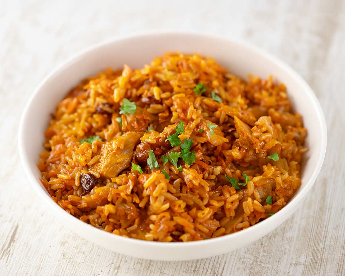 Veganer Plov in einer weißen Schale, zubereitet mit Reis, Karotten, Rosinen und veganem Fleischersatz, garniert mit frischer Petersilie.