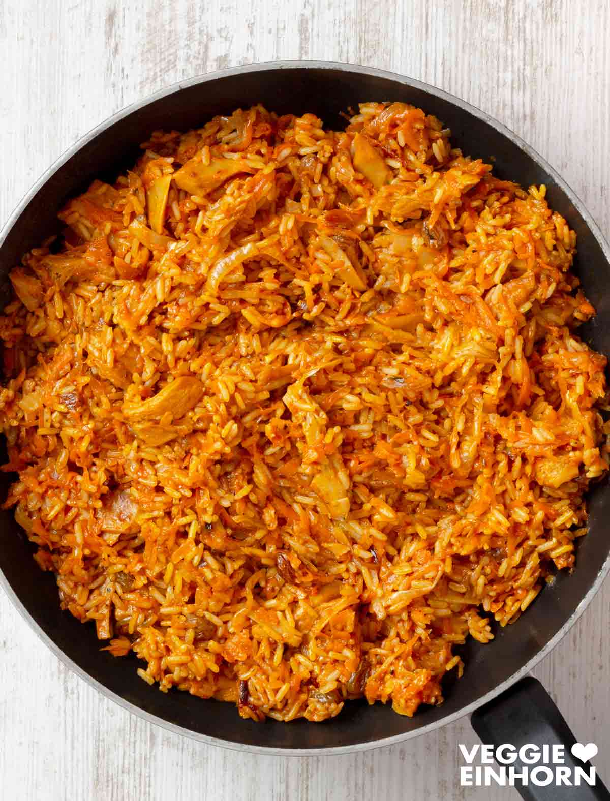 Plov mit veganem Fleisch, Möhren und Rosinen. Veganer Plov in einer schwarzen Pfanne, zubereitet mit Reis, Karotten und veganem Fleischersatz, auf einem hellen Holztisch serviert.