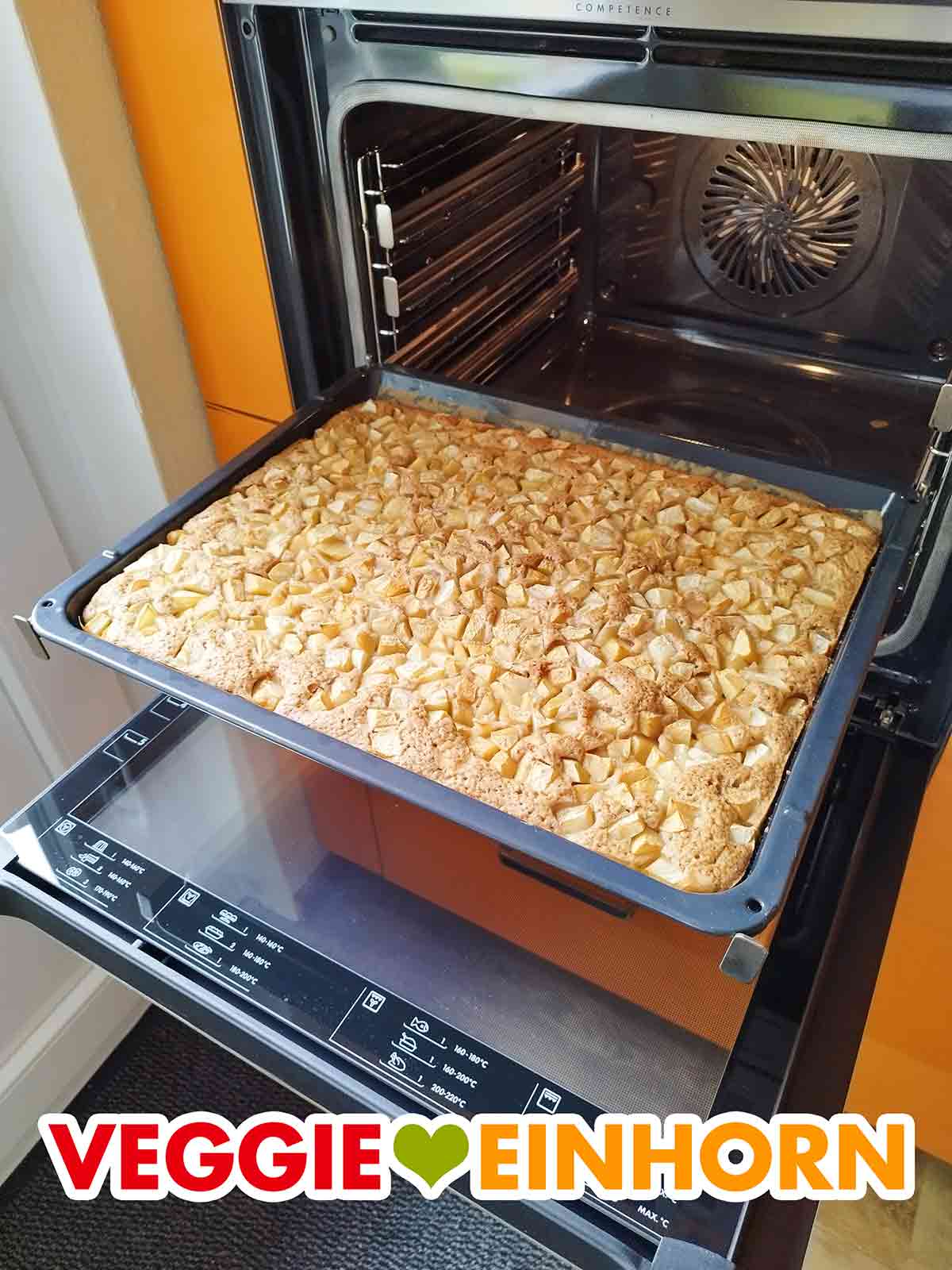 Der vegane Apfelkuchen wird auf dem großen, tiefen Backblech gebacken, das zum Ofen dazu gehört. Geöffneter Backofen mit einem Backblech mit veganem Apfelkuchen