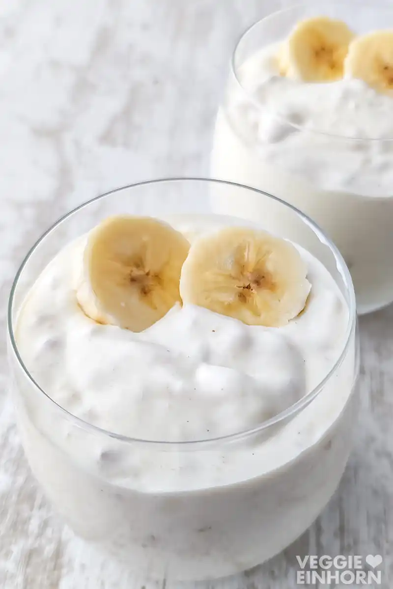 Nahaufnahme eines Glases mit cremigem veganem Bananenjoghurt, dekoriert mit zwei Bananenscheiben. Im Hintergrund ein weiteres Glas.